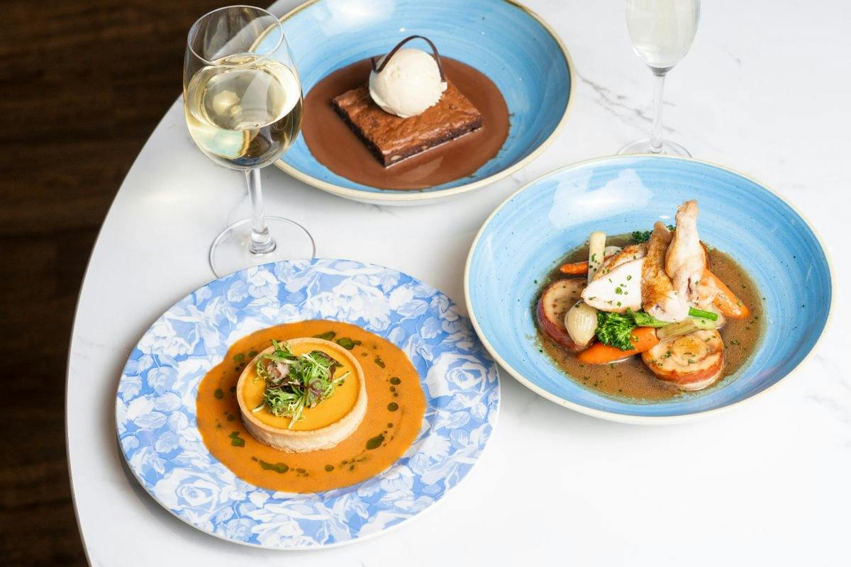 Three gourmet dishes with wine on a white table: a tart, chicken with vegetables, and a brownie with ice cream.