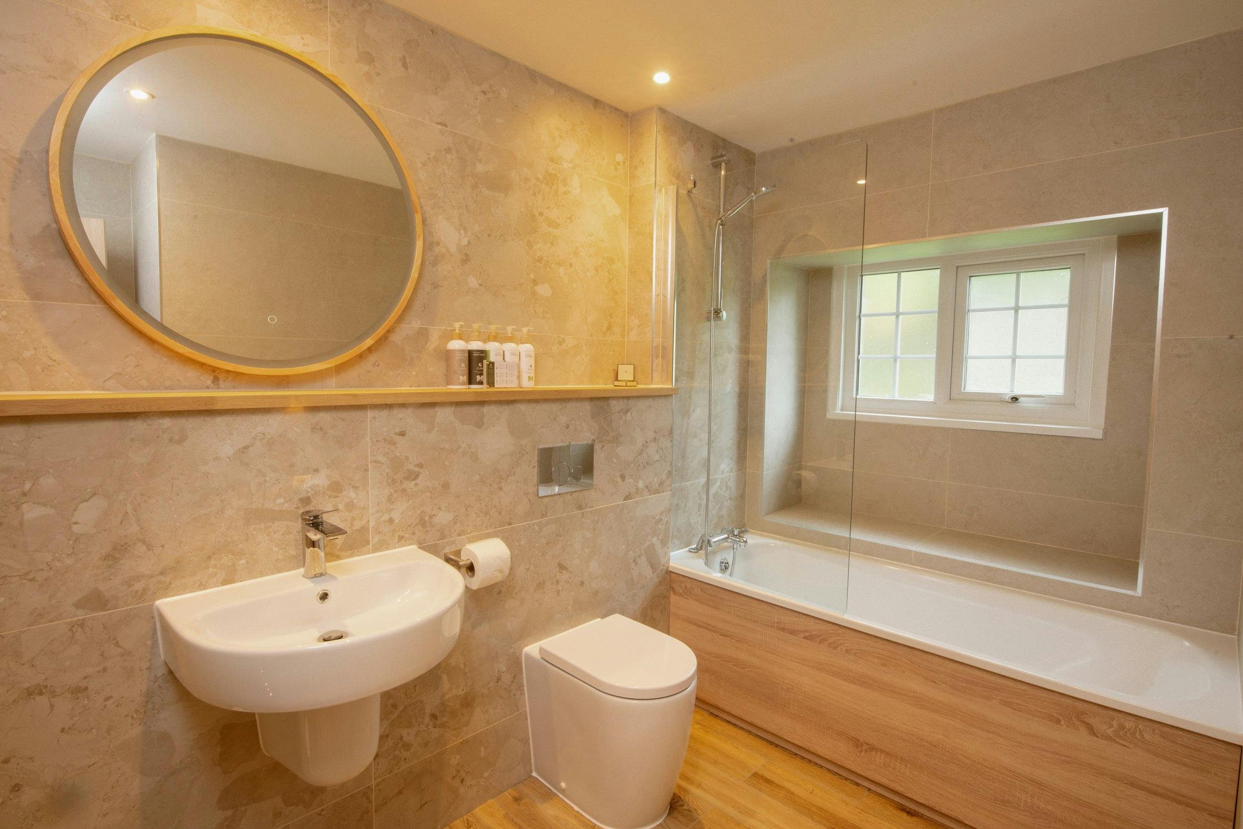 Modern beige-tiled bathroom with round mirror, wall-mounted sink, toilet, and bathtub with glass shower screen beneath a window.