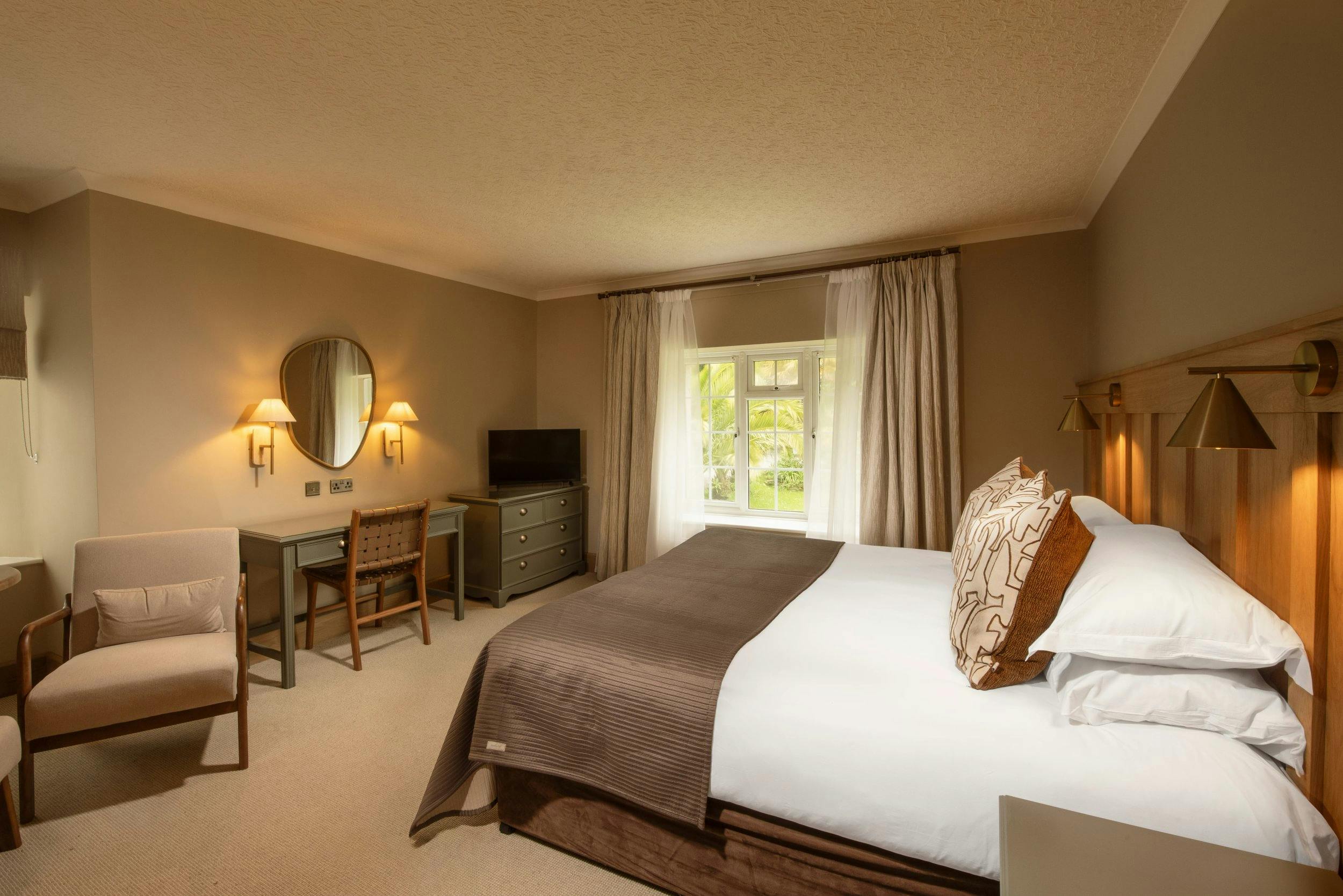 Warm, neutral-toned bedroom with a double bed, wooden headboard, desk, chair, TV, and a window with sheer curtains.