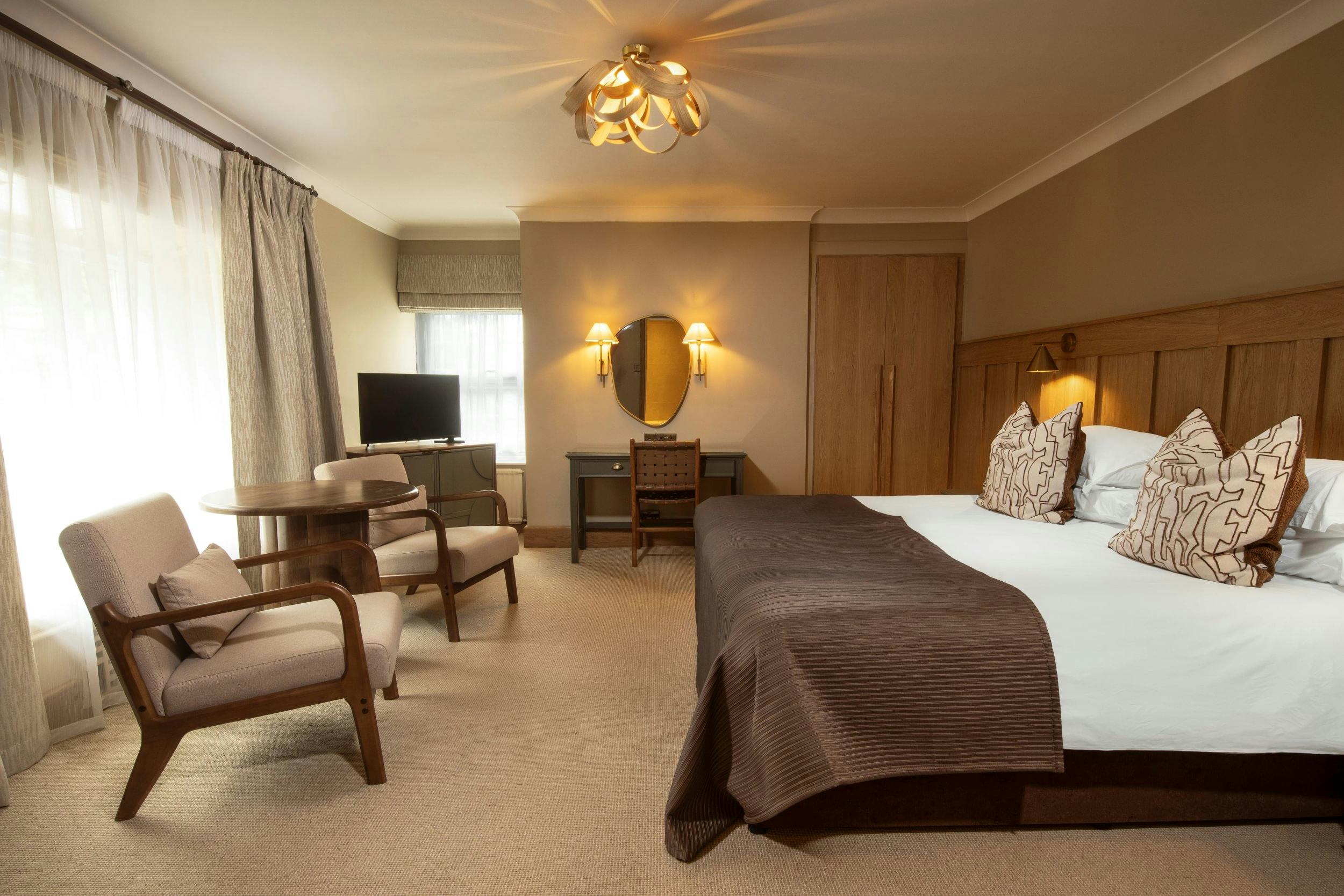 Warm, modern hotel room with a large bed, wooden headboard, two armchairs, round table, TV, desk, and soft window light.