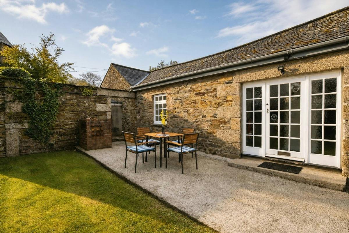 Stone cottage patio with a small table and four chairs, French doors, lawn, and ivy-covered wall under a blue sky.
