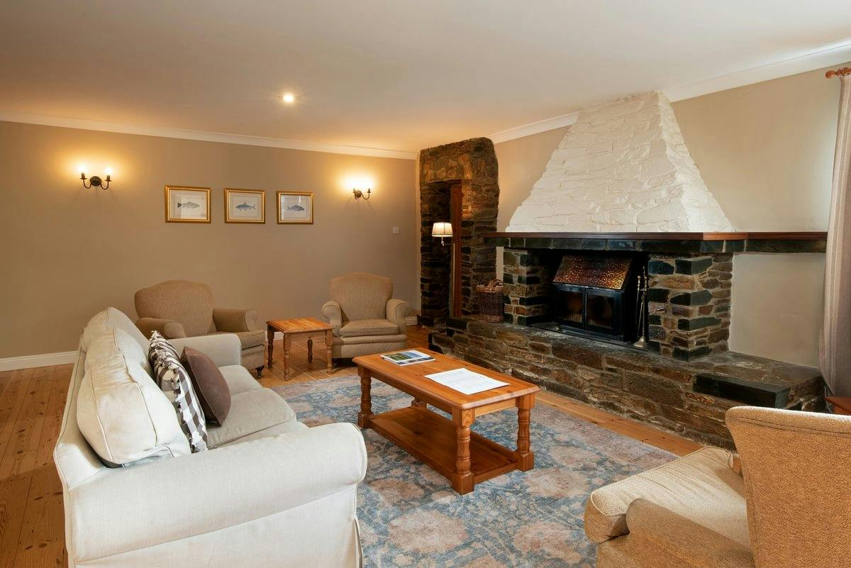 Cosy living room with beige sofas, armchairs, a wooden coffee table, and a large stone fireplace. Wall lights and framed art add warmth.