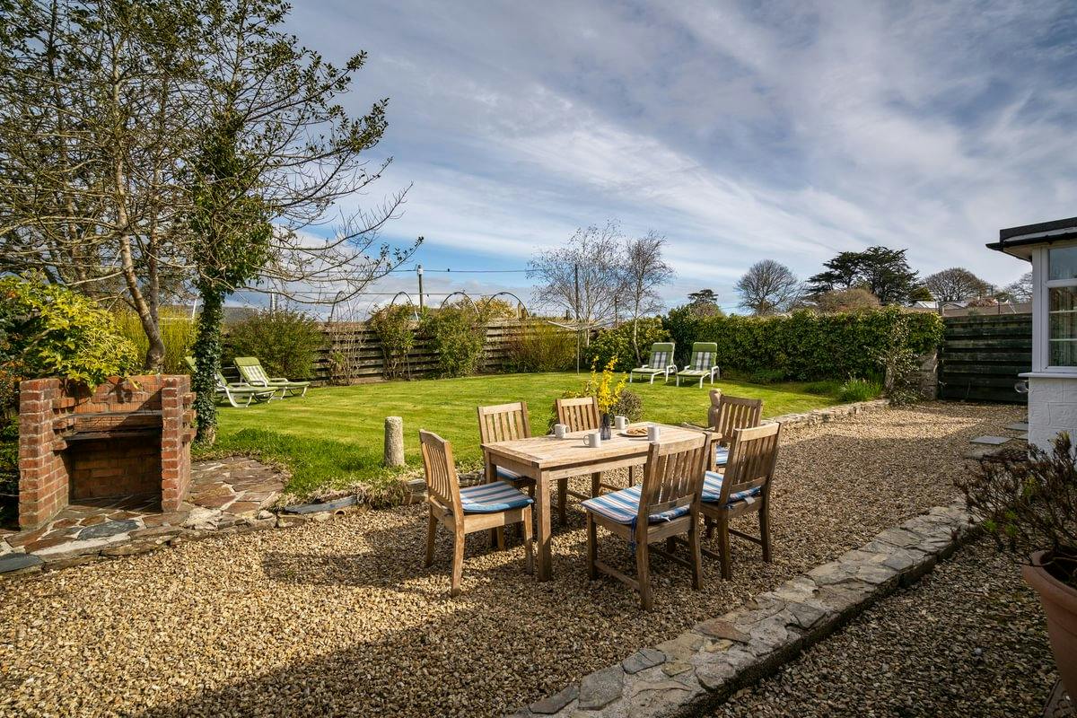 Sunny backyard with gravel patio, wooden dining set, brick barbecue, lawn, trees, and lounge chairs under a streaked sky.