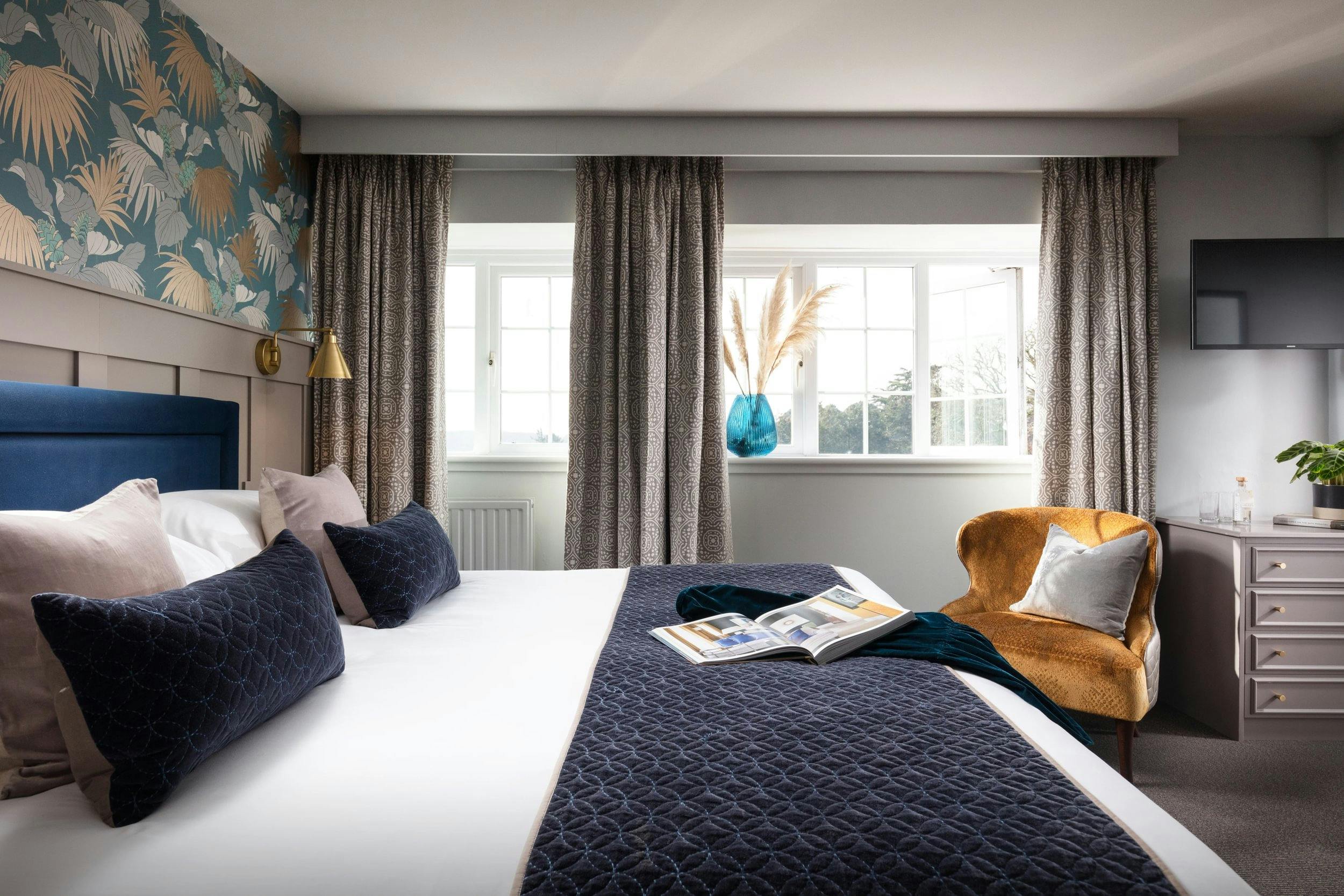 Stylish hotel bedroom with a large bed, blue cushions, patterned curtains, floral wallpaper, and a mustard armchair by bright windows.
