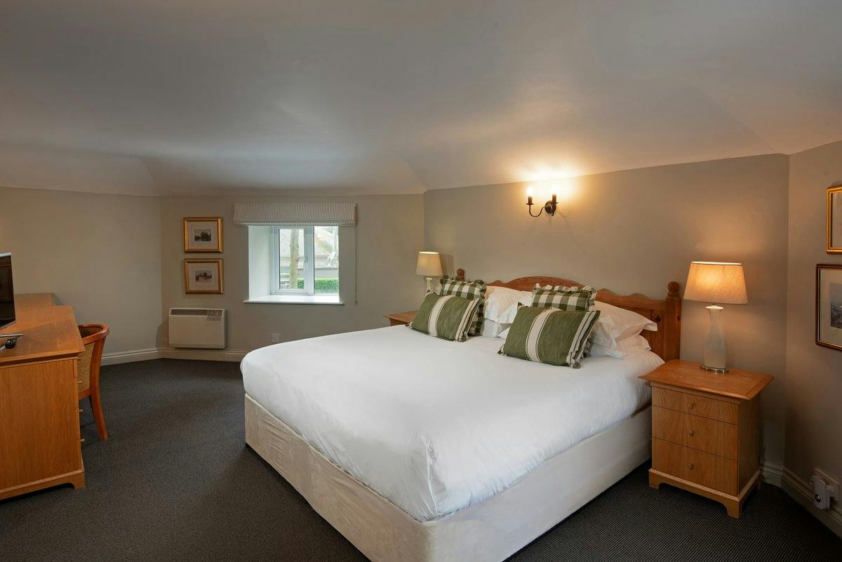Cosy hotel room with a large bed, green pillows, wooden furniture, lamps, and a small window.