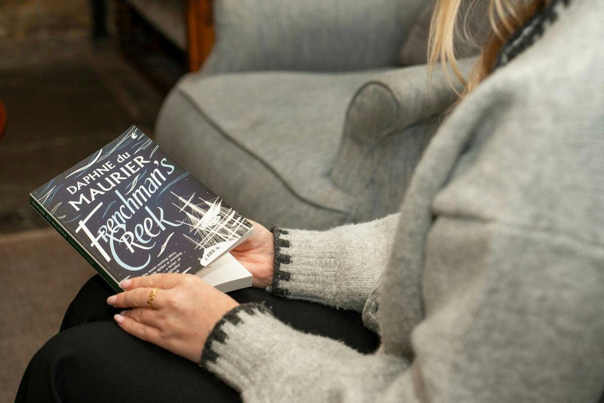 Person in a gray sweater sits on a couch holding a copy of Daphne du Maurier’s Frenchman’s Creek.