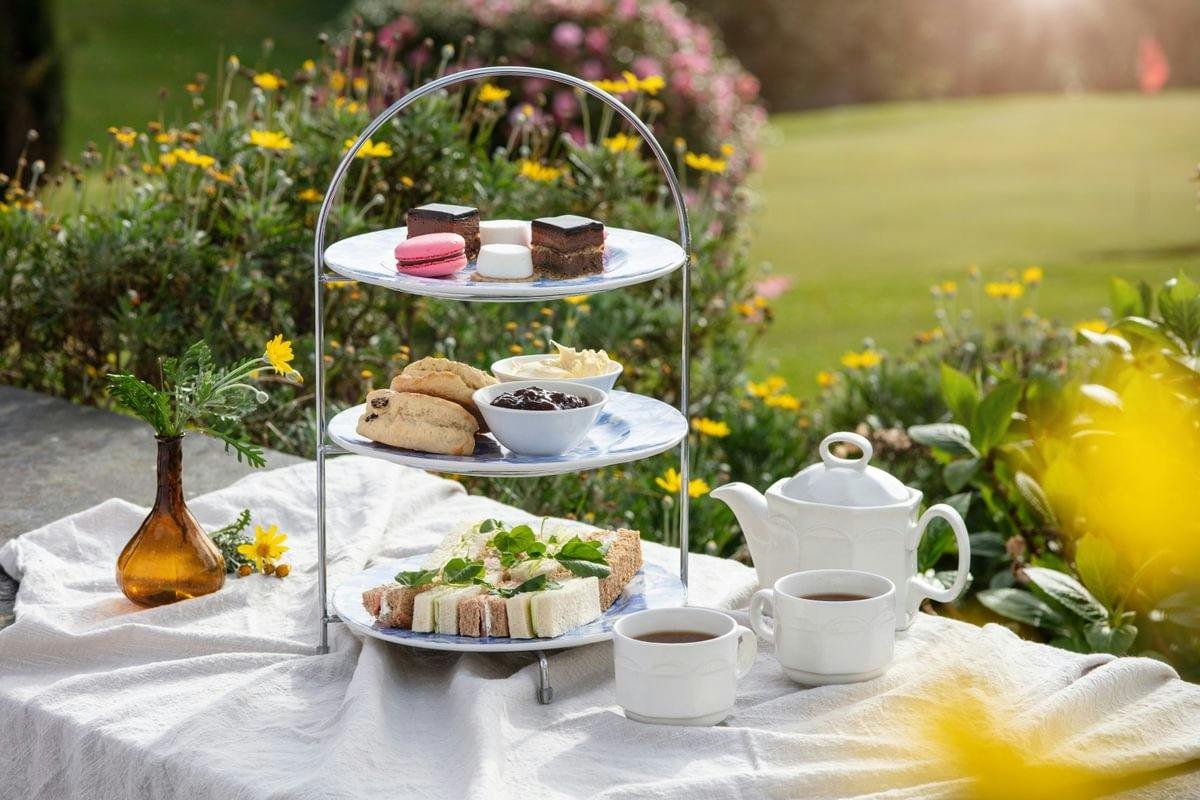Afternoon tea setup outdoors with a tiered tray of sandwiches, scones, and desserts, plus a teapot and cups on a white cloth.