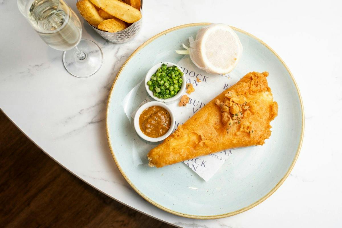 Plate with battered fish, mushy peas, curry sauce, lemon slice, chips in a basket, and a glass of white wine on a marble table.