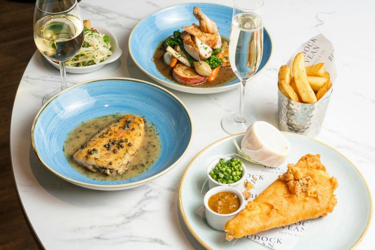 A table with fish dishes, fries, peas, sauces, and glasses of white wine and champagne on a marble surface.