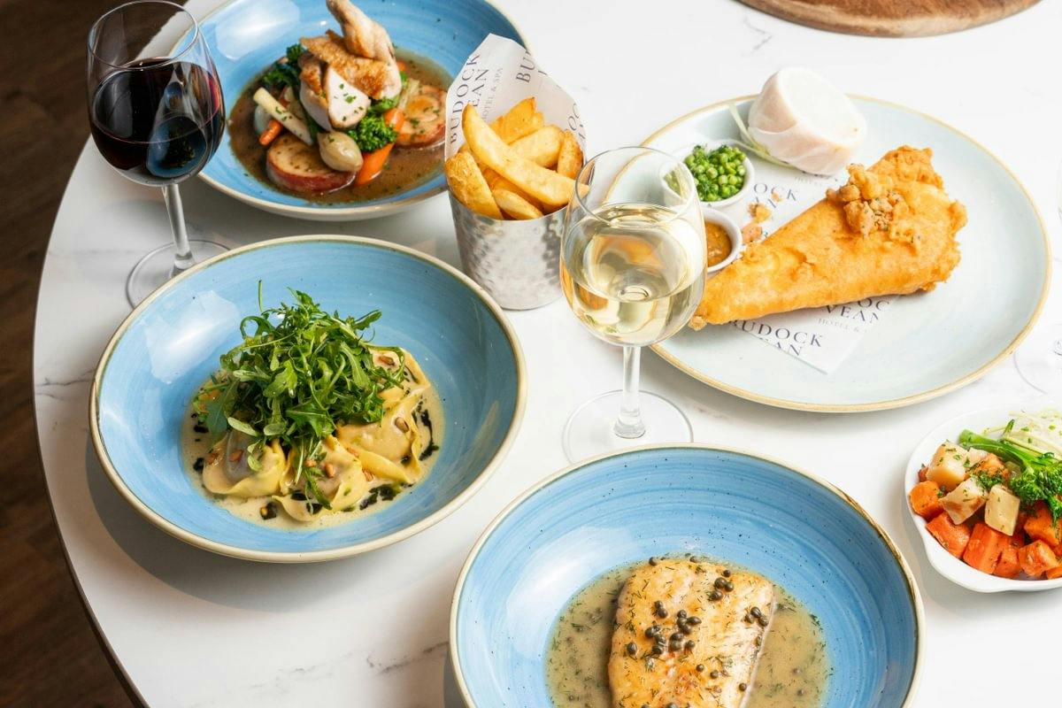 A table with fish and chips, pasta, chicken, vegetables, red and white wine.
