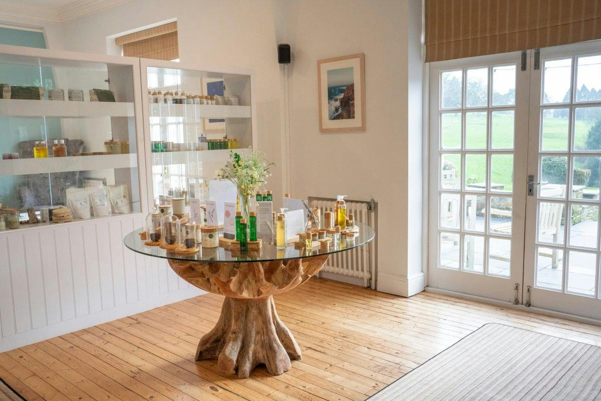 A bright room with a wooden floor, glass table displaying skincare products, shelves with more items, and large windows overlooking greenery.
