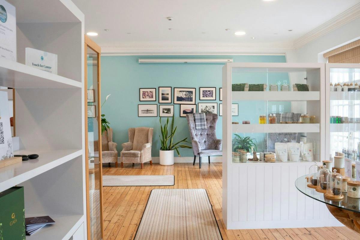 Bright room with teal walls, cosy chairs, plants, and shelves displaying products. Framed photos adorn the wall.