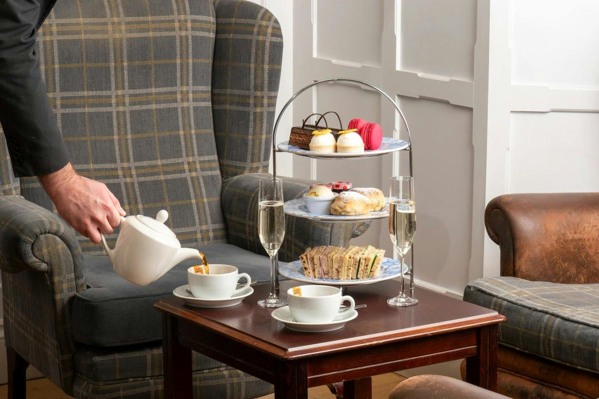 A person pours tea into a cup beside a tiered tray of pastries and sandwiches, with two glasses of champagne on a table.