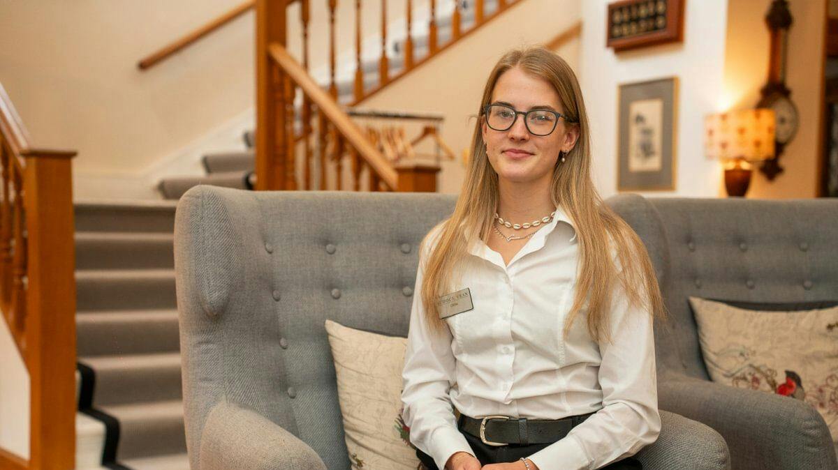 Woman in glasses and white shirt sits on a grey chair in a cosy room with wooden stairs and framed art.