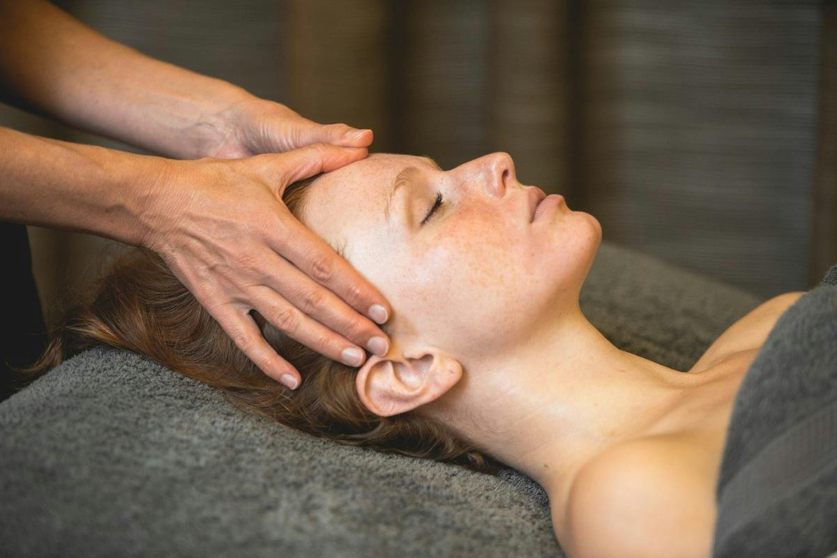 Person lying on a spa table with eyes closed while another person gently massages their head and temples.
