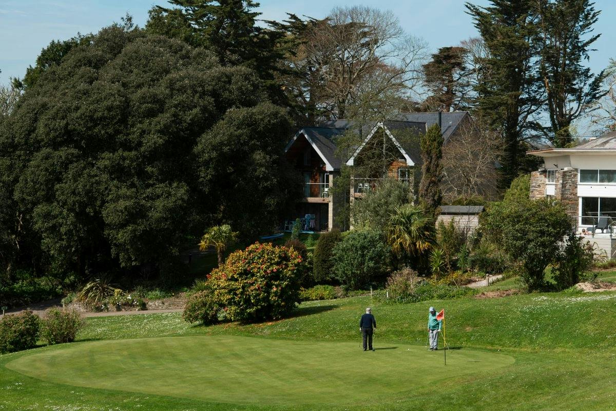 Two golfers stand on a putting green beside a flag, with houses, shrubs, and tall trees in the background.