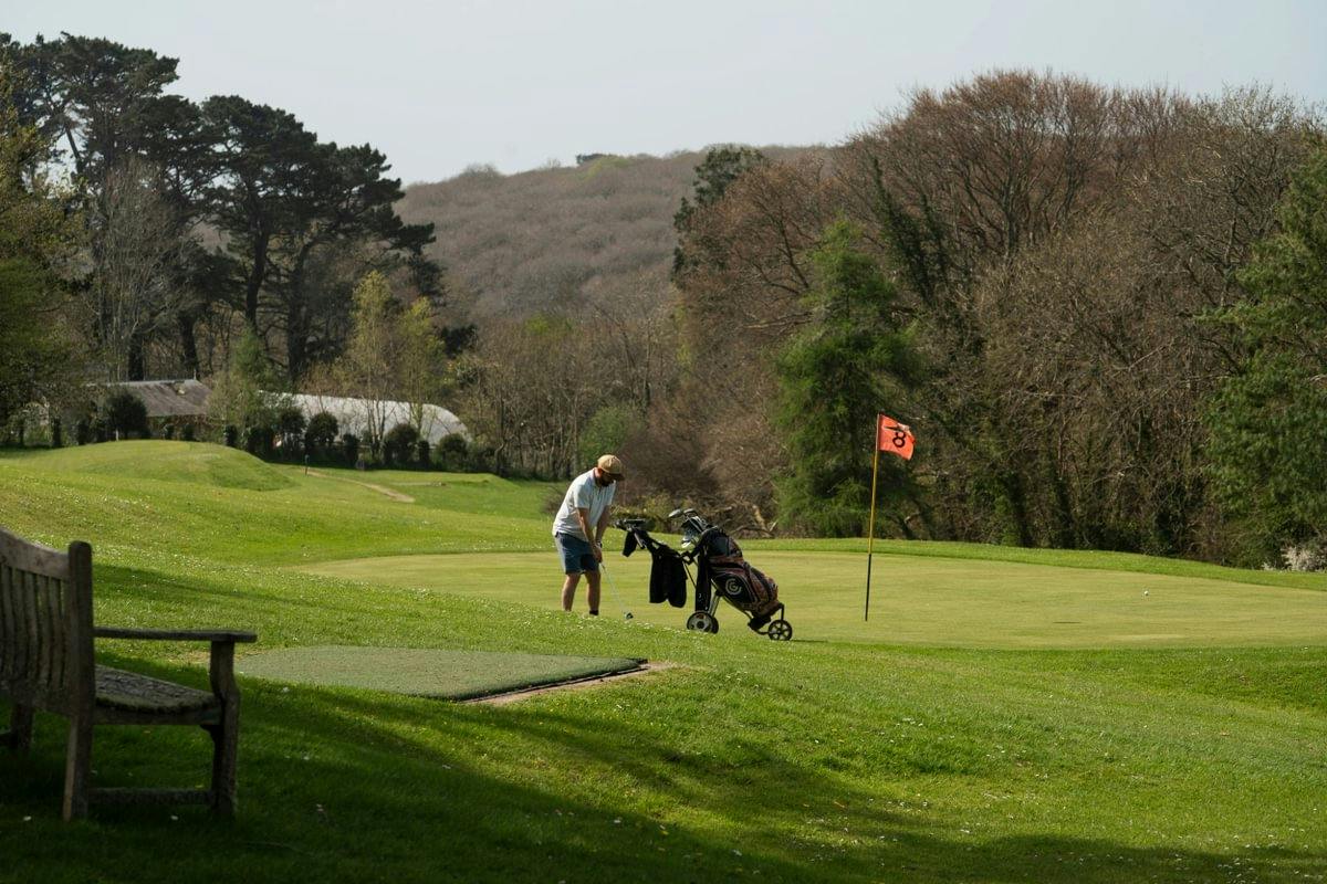 Golfer putts on a green beside a flag and golf cart, with a bench, trees, and rolling hills in the background.