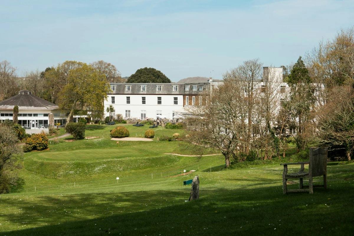 Large white hotel overlooking a manicured lawn and putting green, with trees, shrubs, and a bench under a clear sky.