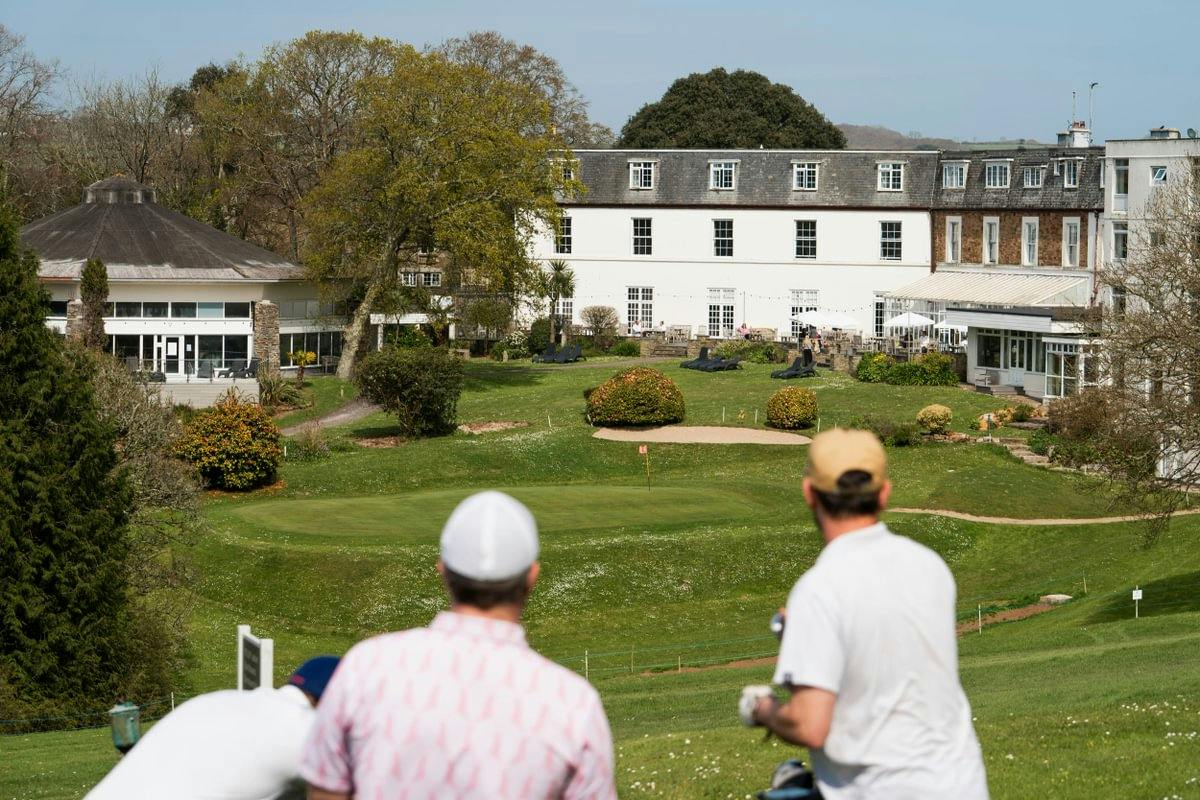 Golfers on a hill overlook a putting green and sand bunker in front of a large hotel and landscaped gardens.