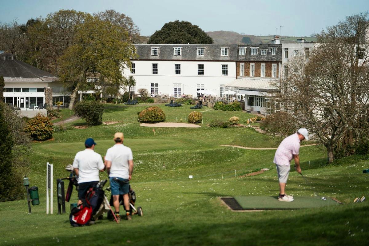 Golfer tees off on a green beside two players with golf bags, with a large hotel and trees in the background.