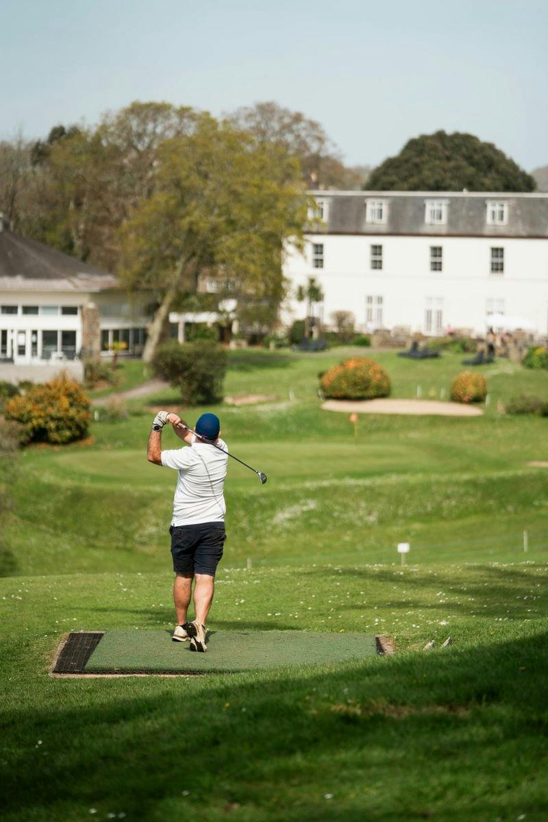 Golfer in a white shirt follows through on a tee shot toward a green, with a large white clubhouse in the background.
