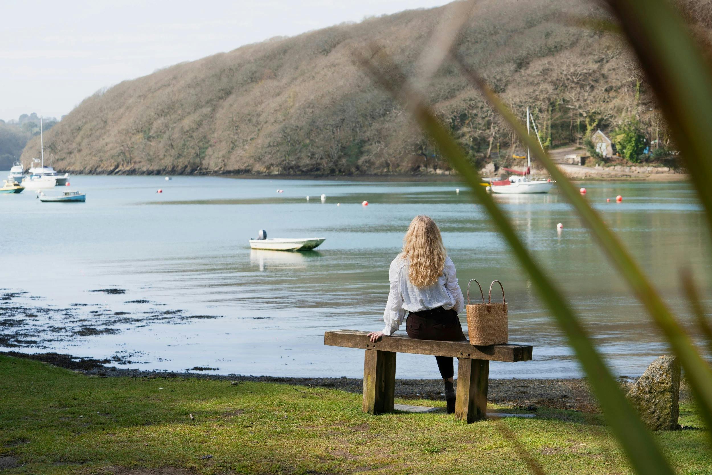 Person with long blond hair sits on a bench by a calm estuary, looking at small boats on the water, with a woven bag beside them.
