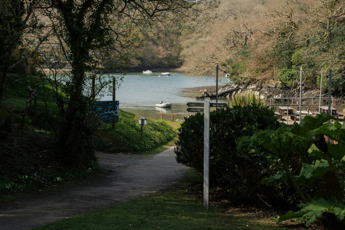 Shaded path leads to a quiet riverside cove with small boats, trees, and a signpost near a seating area.