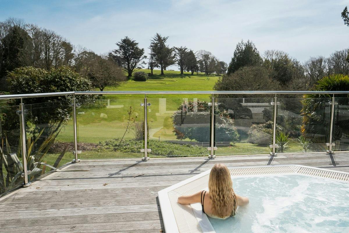 Person relaxing in an outdoor hot tub on a wooden deck, overlooking a green lawn and trees beyond a glass railing.