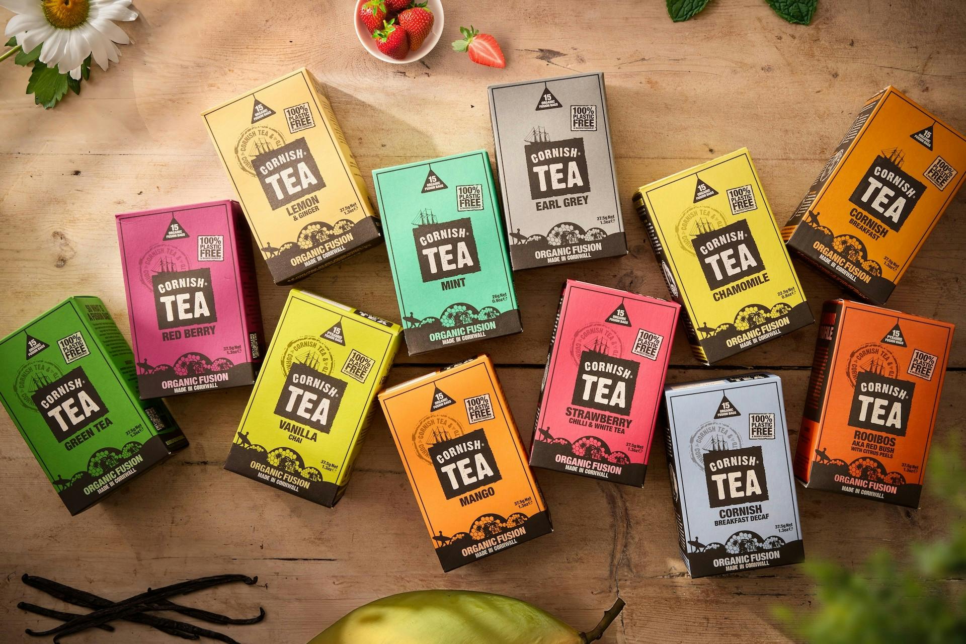 Colourful Cornish Tea boxes in assorted flavours spread across a wooden table, with strawberries, mint, vanilla pods, and a flower.