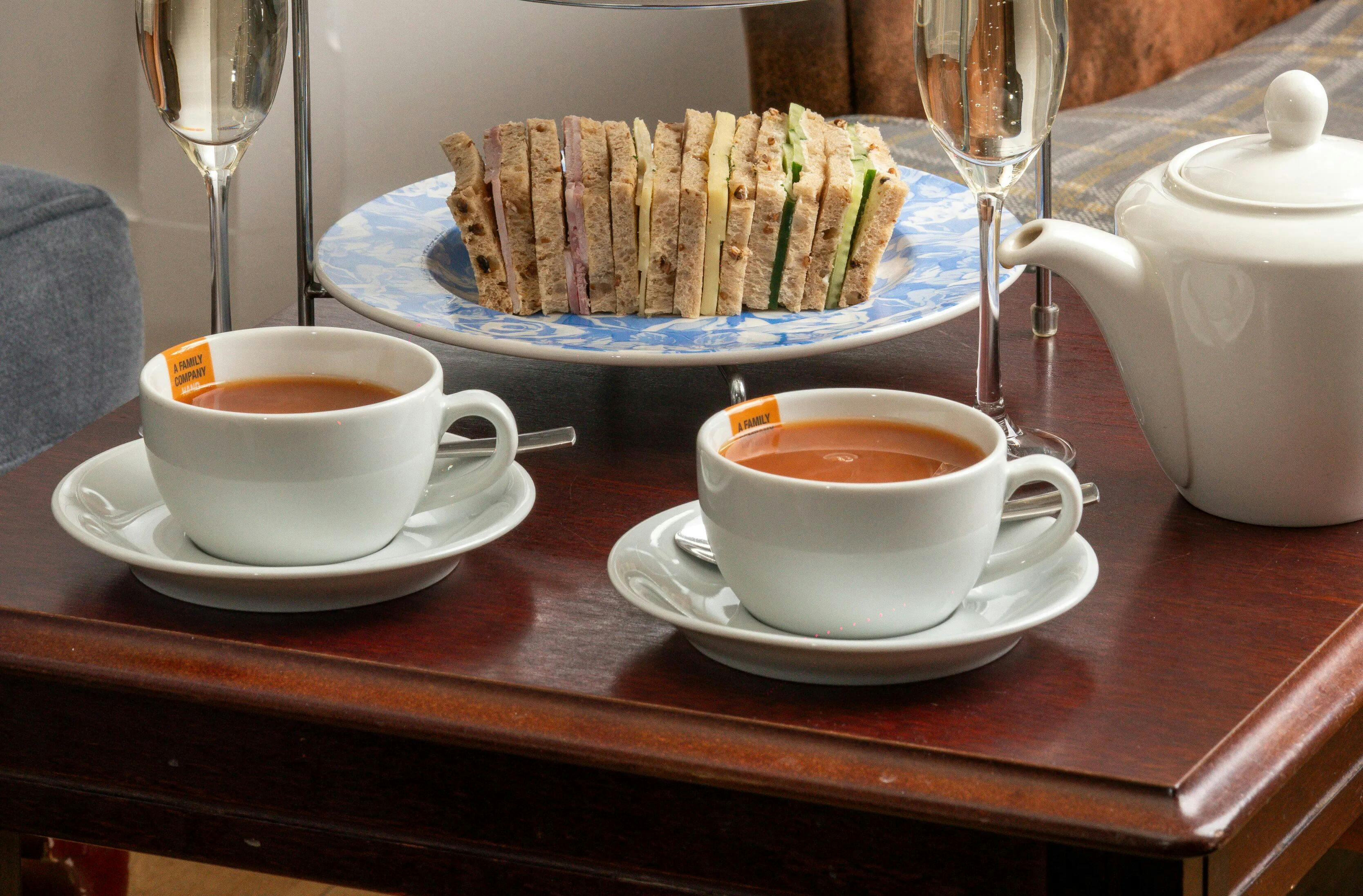 Afternoon tea setup with two cups of tea, a teapot, and assorted finger sandwiches on a tiered stand.