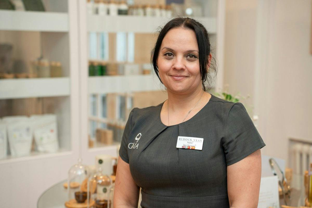 Smiling spa staff member in a dark uniform with a name badge, standing in a bright treatment room with shelves of products behind her.