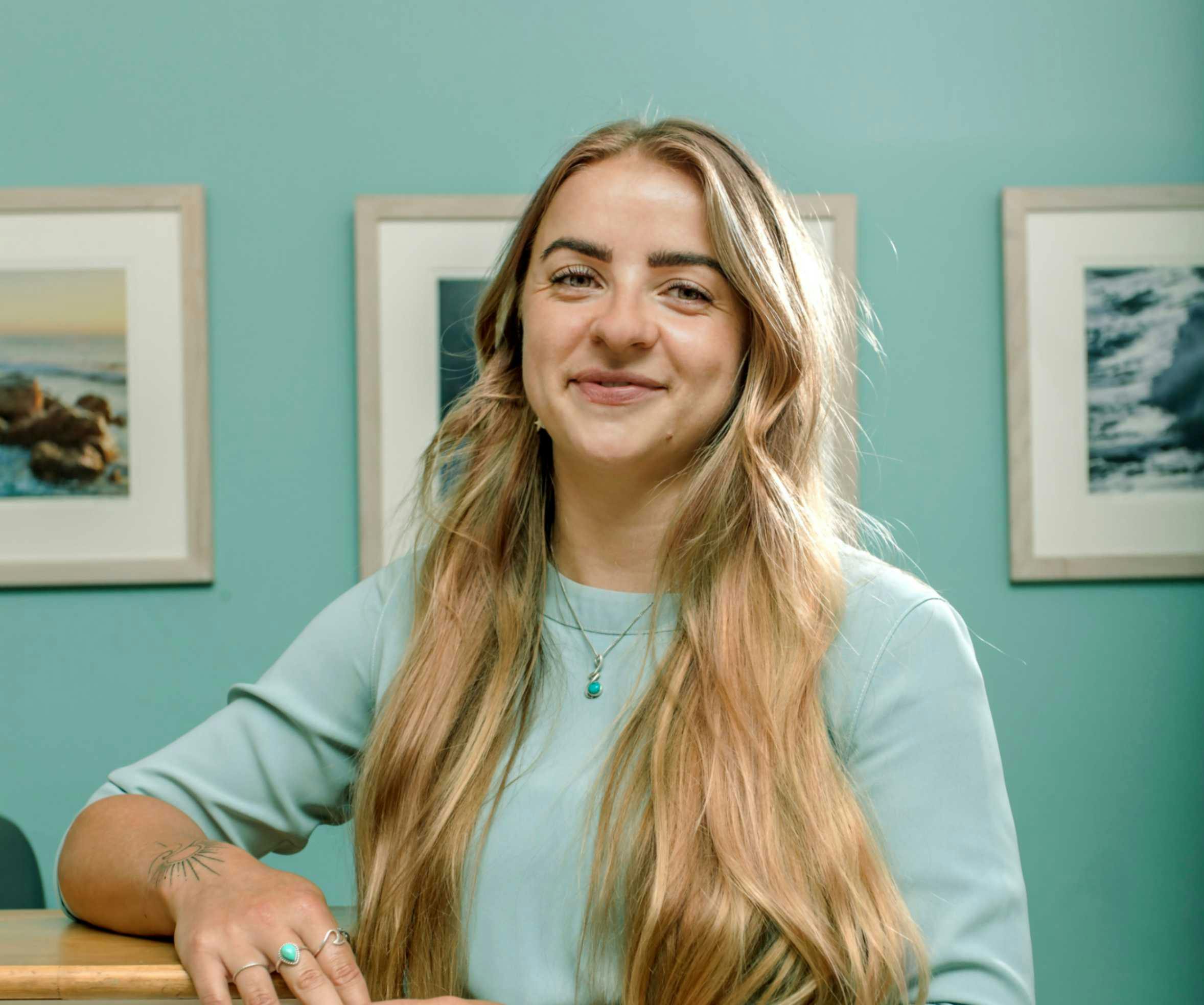 Smiling woman with long blonde hair in a light blue top, leaning on a desk against a teal wall with framed ocean photos.