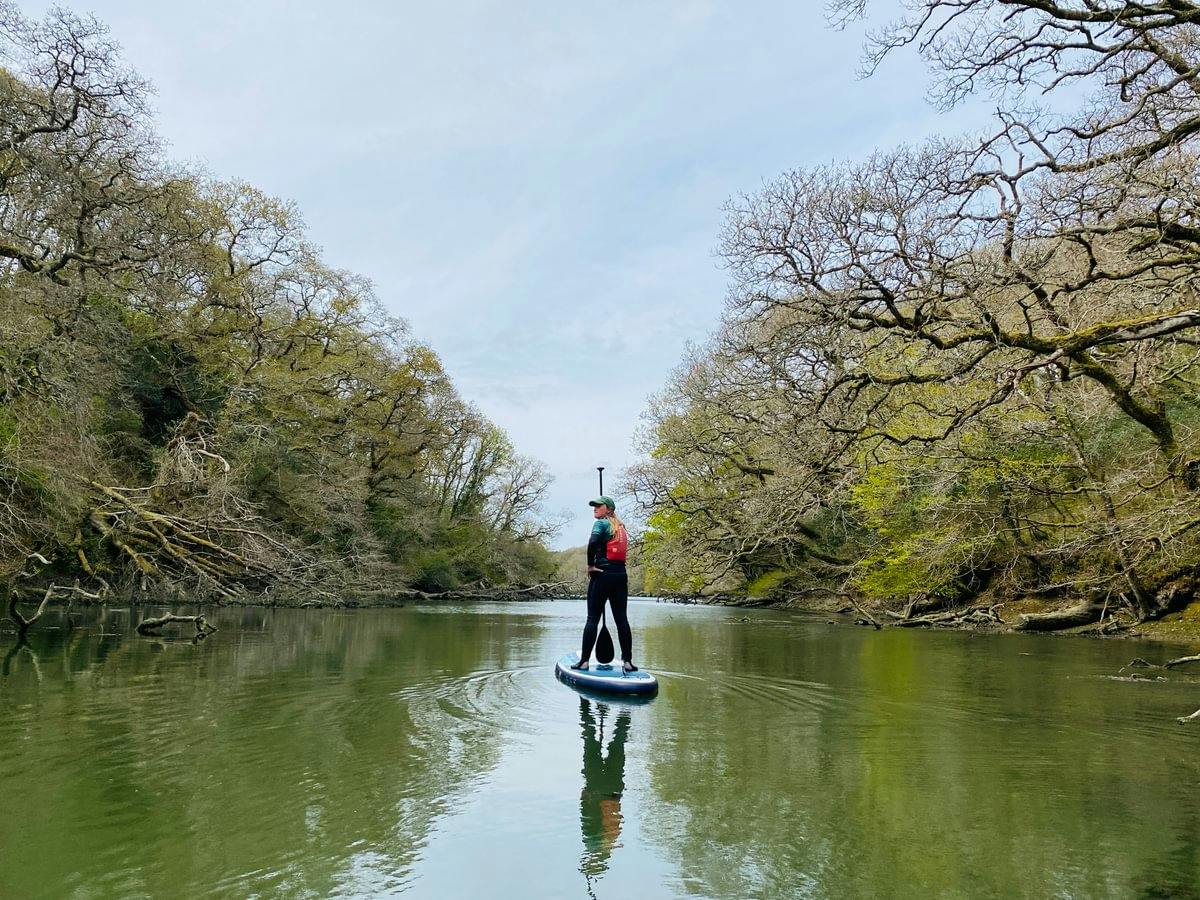 Person paddleboarding on a calm, tree-lined river, reflected in the green water beneath a pale blue sky.