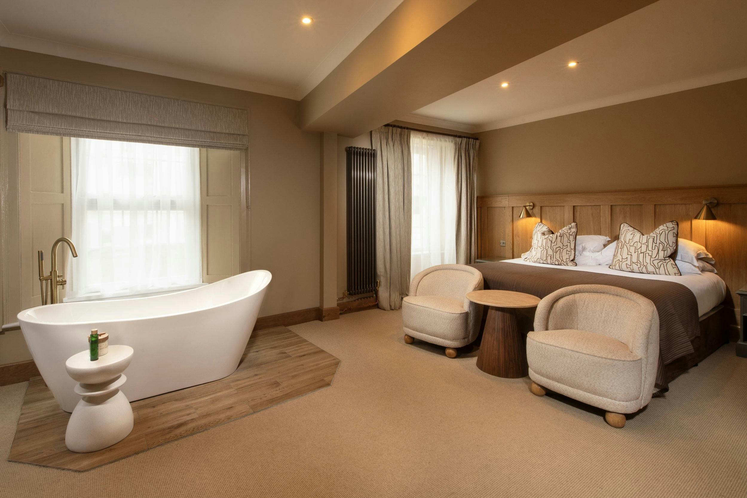 Warm, modern bedroom with a king bed, two armchairs, and a freestanding bathtub by a large curtained window.