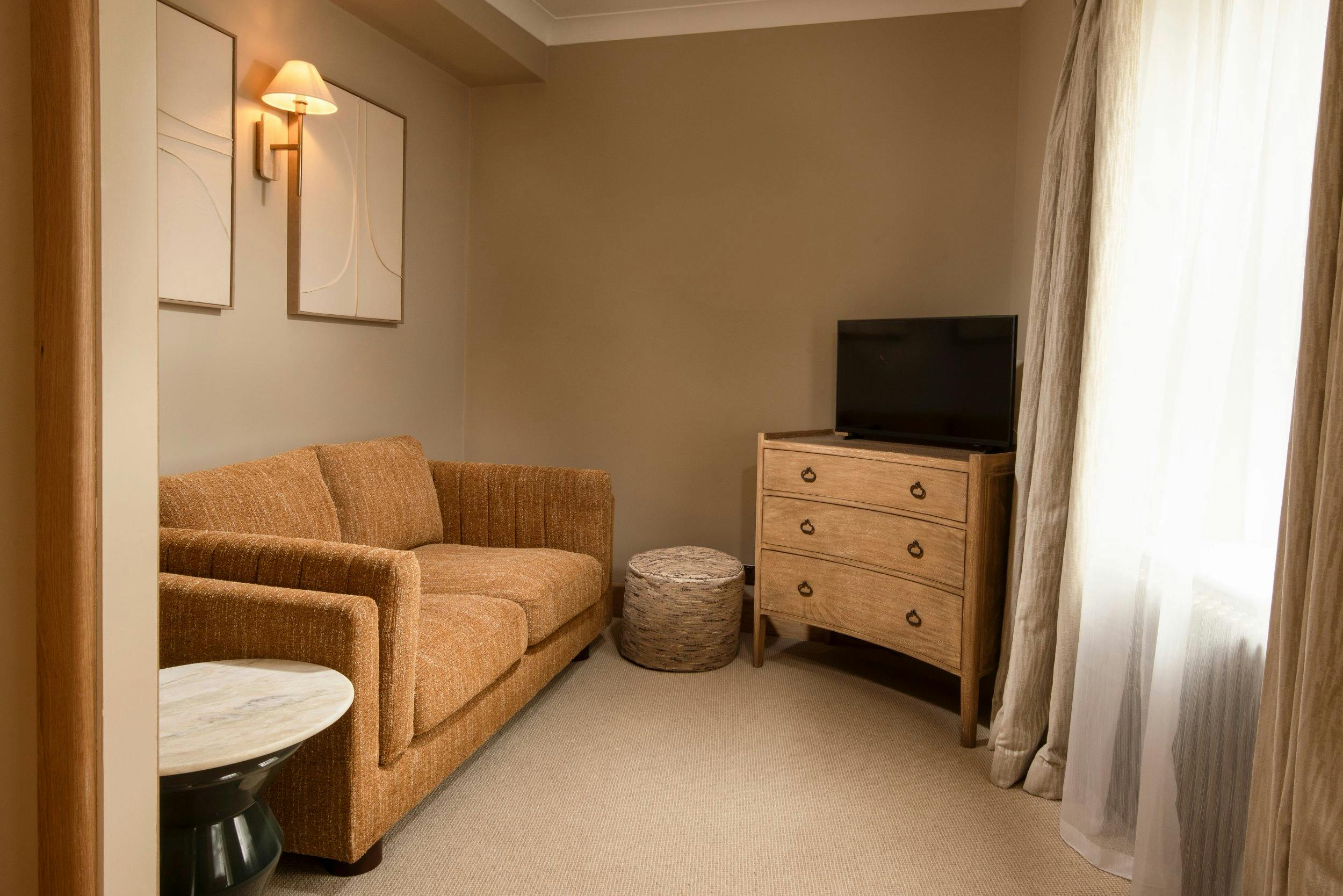 Cosy sitting area with a brown loveseat, wall lamp, abstract art, TV on a wooden dresser, and sheer-curtained window.
