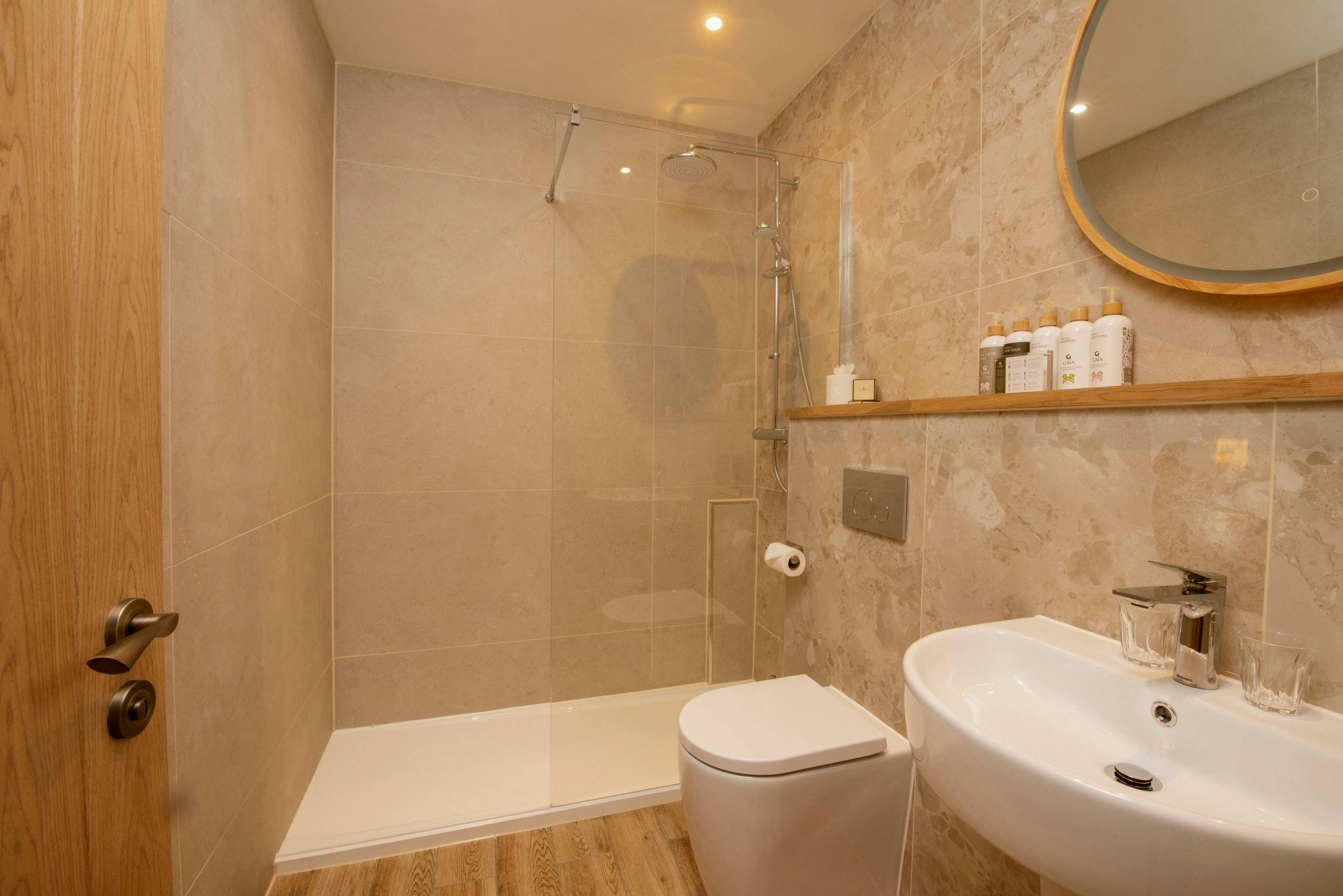 Modern beige-tiled bathroom with walk-in glass shower, toilet, pedestal sink, wood shelf, and round mirror.