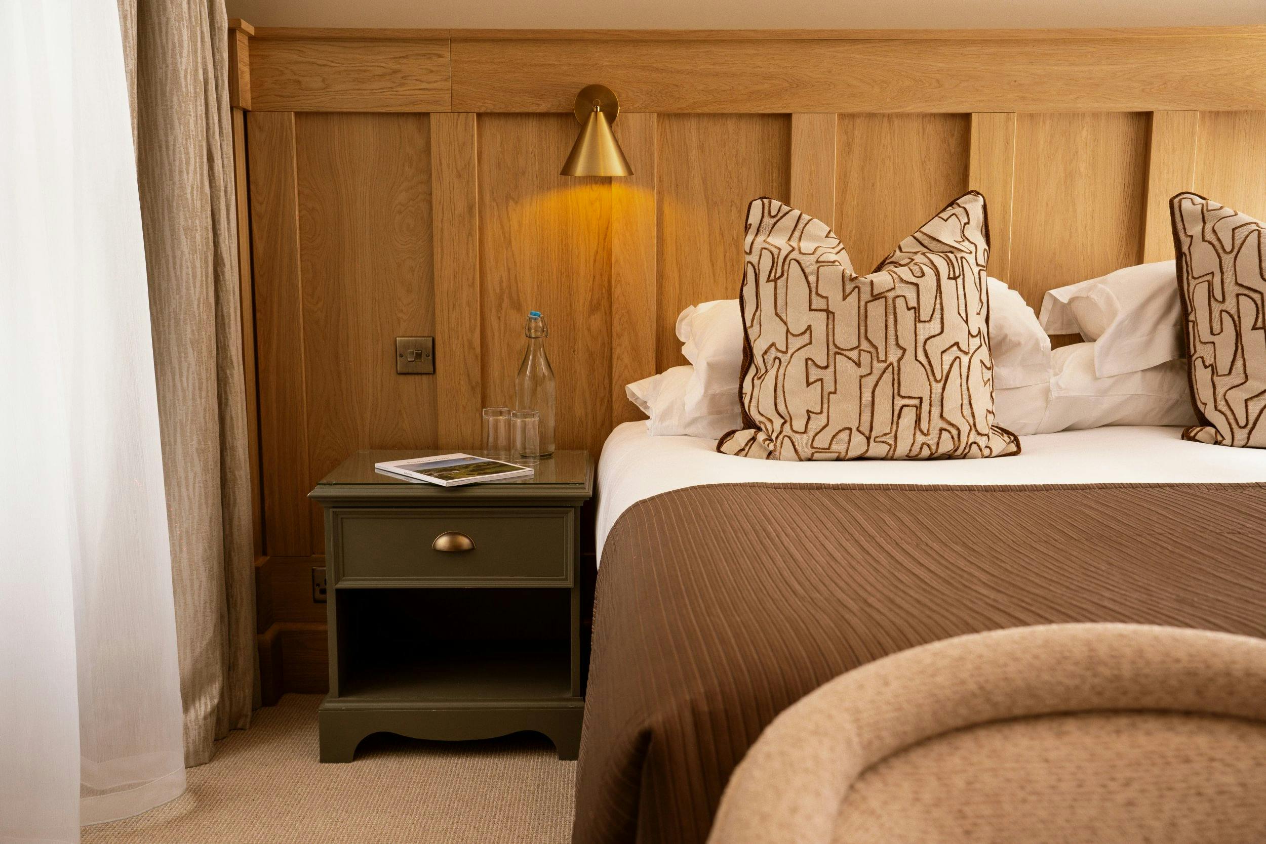 Warm hotel bedroom with wood-paneled headboard, brass reading light, bedside table, and neatly made bed with patterned pillows.