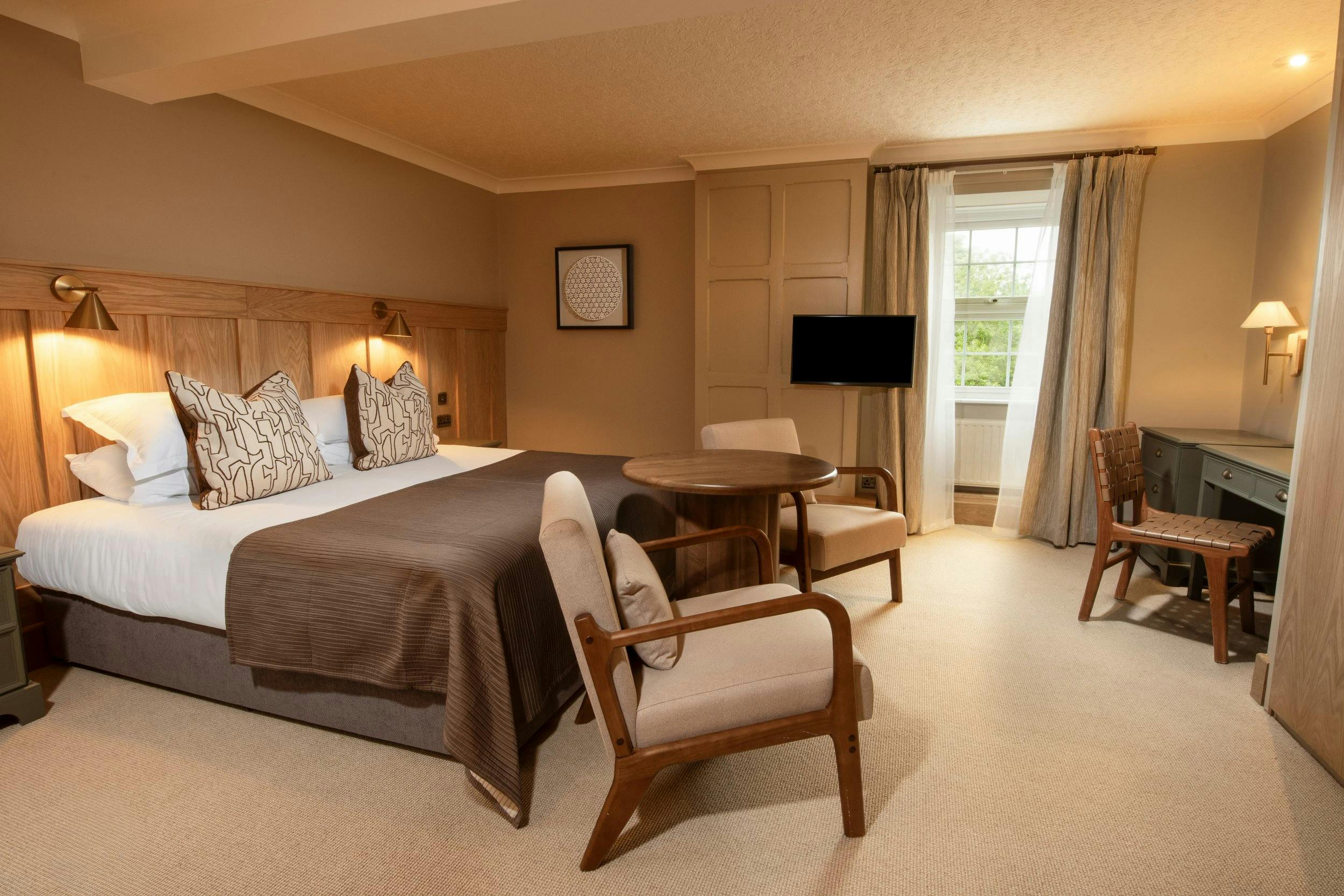 Warm, modern hotel room with a double bed, two armchairs and table, desk, TV, and window with curtains.