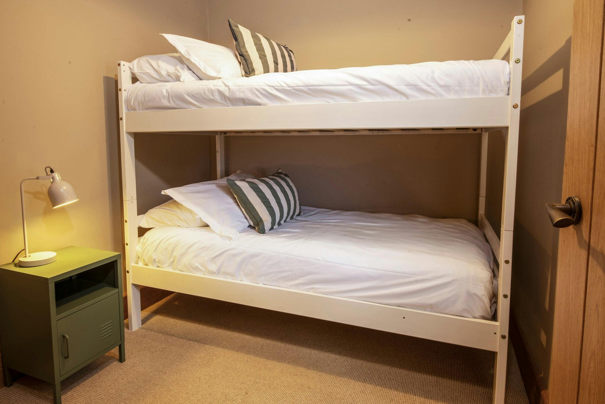 White bunk bed with neatly made white bedding, striped pillows, and a lit bedside lamp on a small green nightstand.