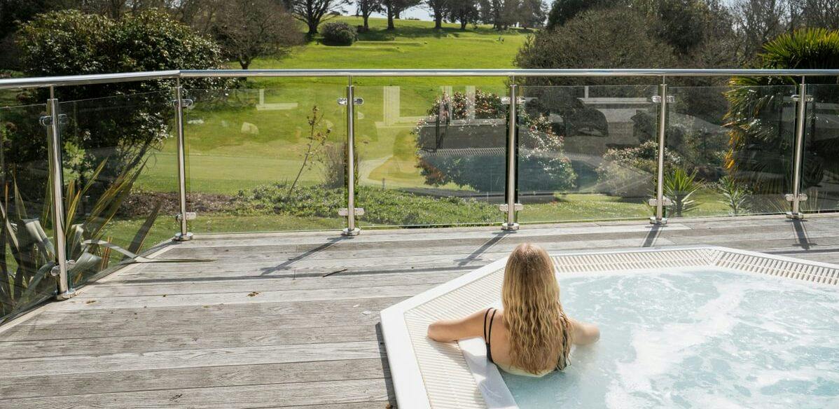 Person relaxing in an outdoor hot tub on a wooden deck, overlooking a green lawn and trees behind a glass railing.