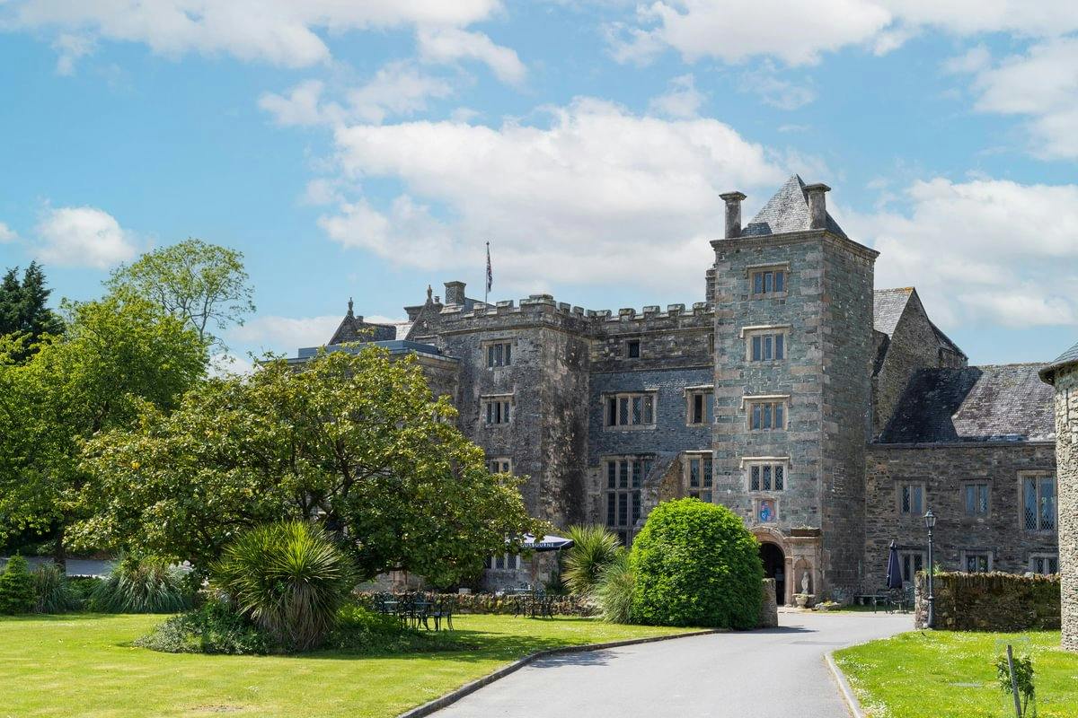 Stone castle-like manor with towers, large trees and lawn, under a bright blue sky with scattered clouds.