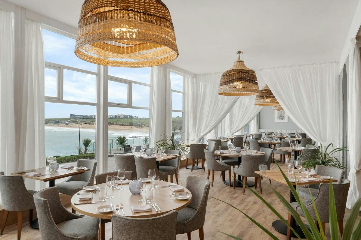 Bright seaside restaurant with wicker pendant lights, white drapes, round tables, gray chairs, and large windows overlooking a beach.