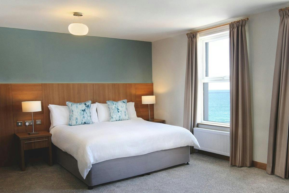 Modern bedroom with a king bed, wooden headboard, bedside lamps, and a window overlooking the sea.