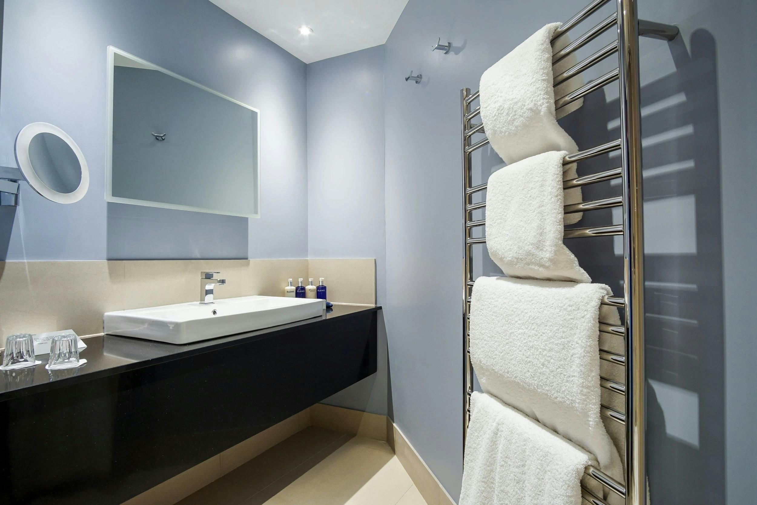 Modern bathroom with a wall-mounted sink, large mirror, black counter, and chrome heated towel rack holding white towels.