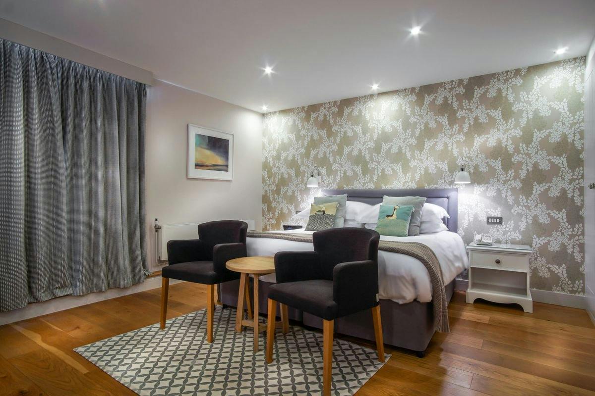 Modern bedroom with floral accent wall, double bed, two dark armchairs, small round table, patterned rug, and floor-to-ceiling curtains.