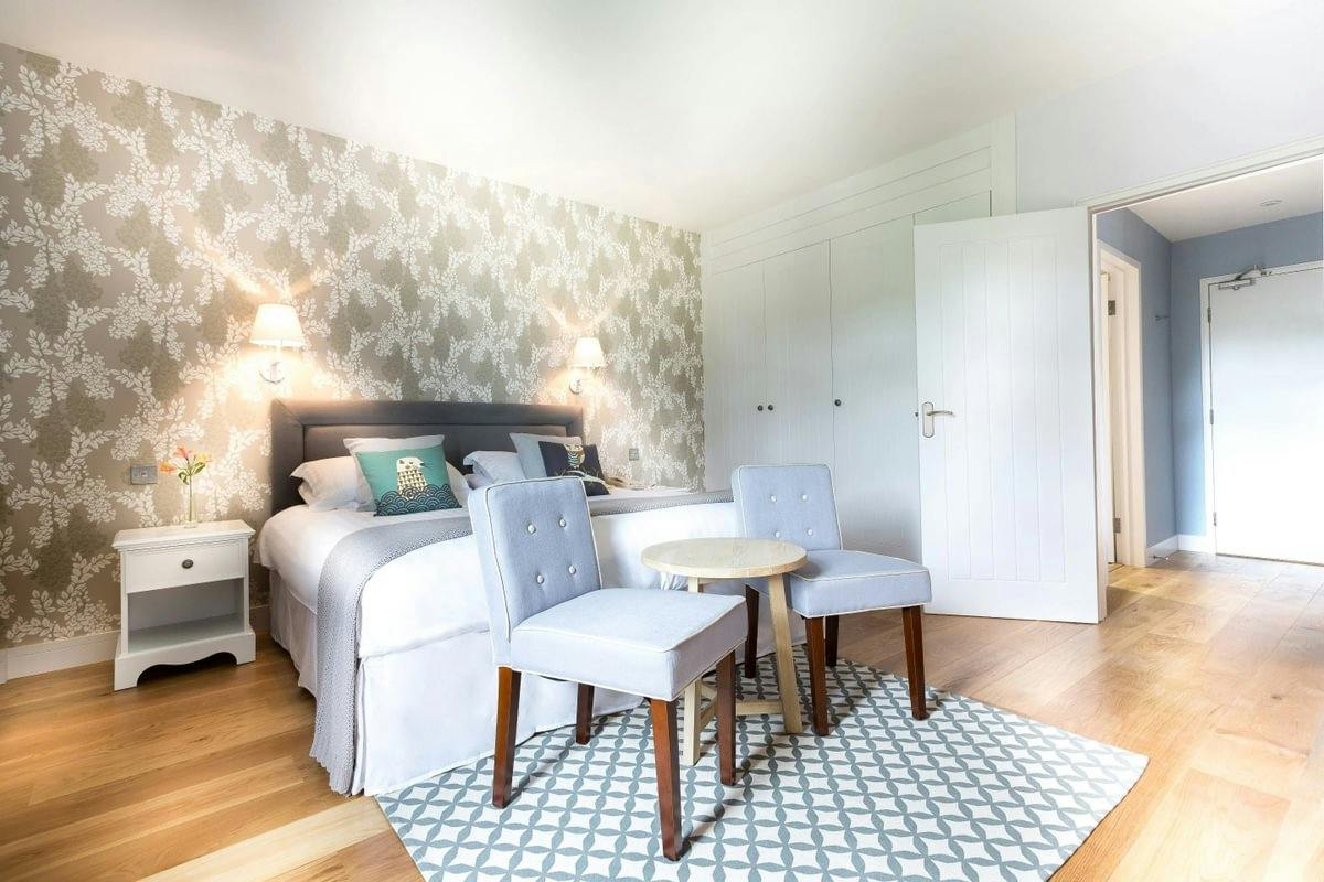 Bright bedroom with floral wallpaper, double bed, bedside table, two upholstered chairs, small round table, and wood floors.