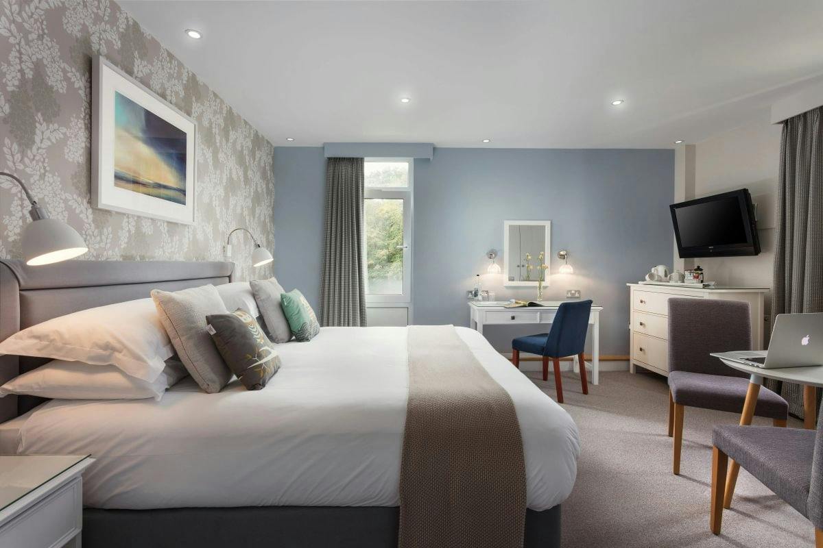 Bright modern hotel room with a large bed, desk and chair, dresser with TV, and soft grey-blue décor.