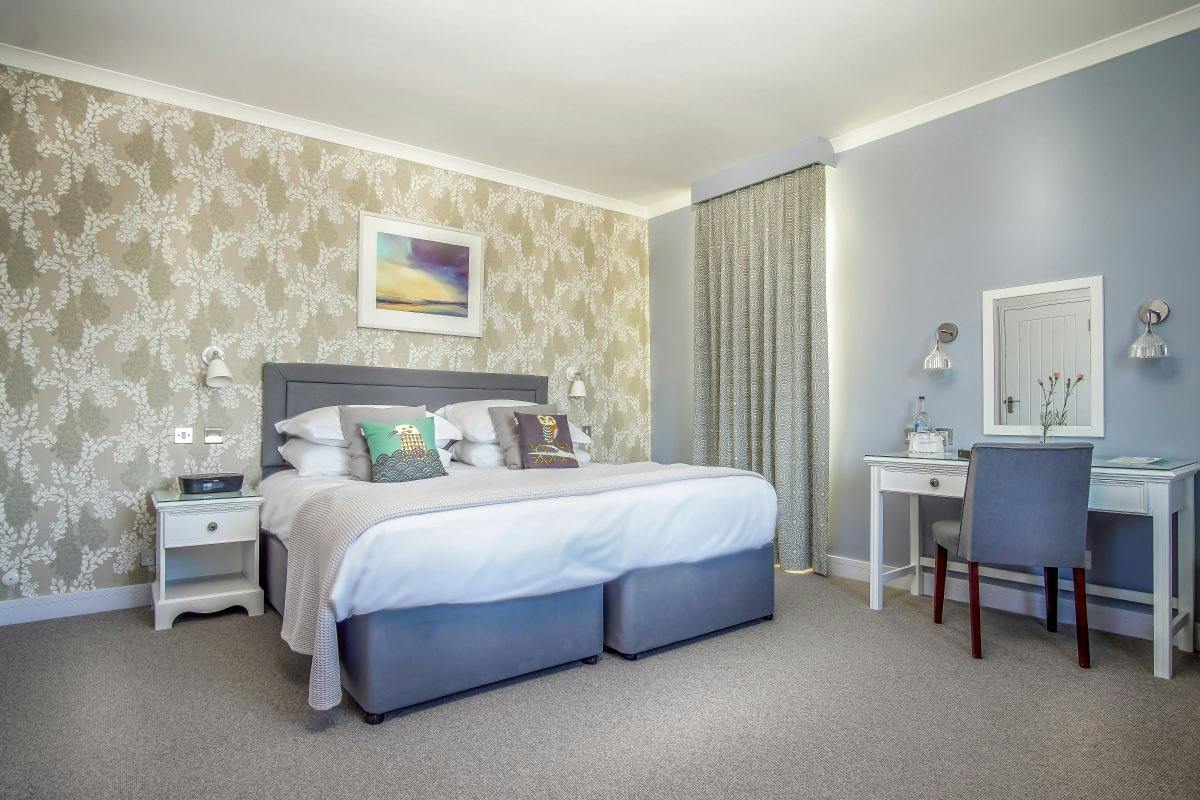 Bright bedroom with a gray upholstered bed, floral accent wall, white nightstand, and vanity desk with chair and mirror.