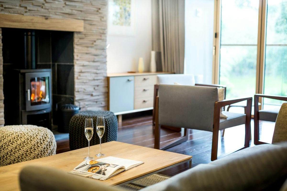 Cosy modern living room with a lit wood stove, stone fireplace, armchairs, and two champagne glasses on a coffee table.