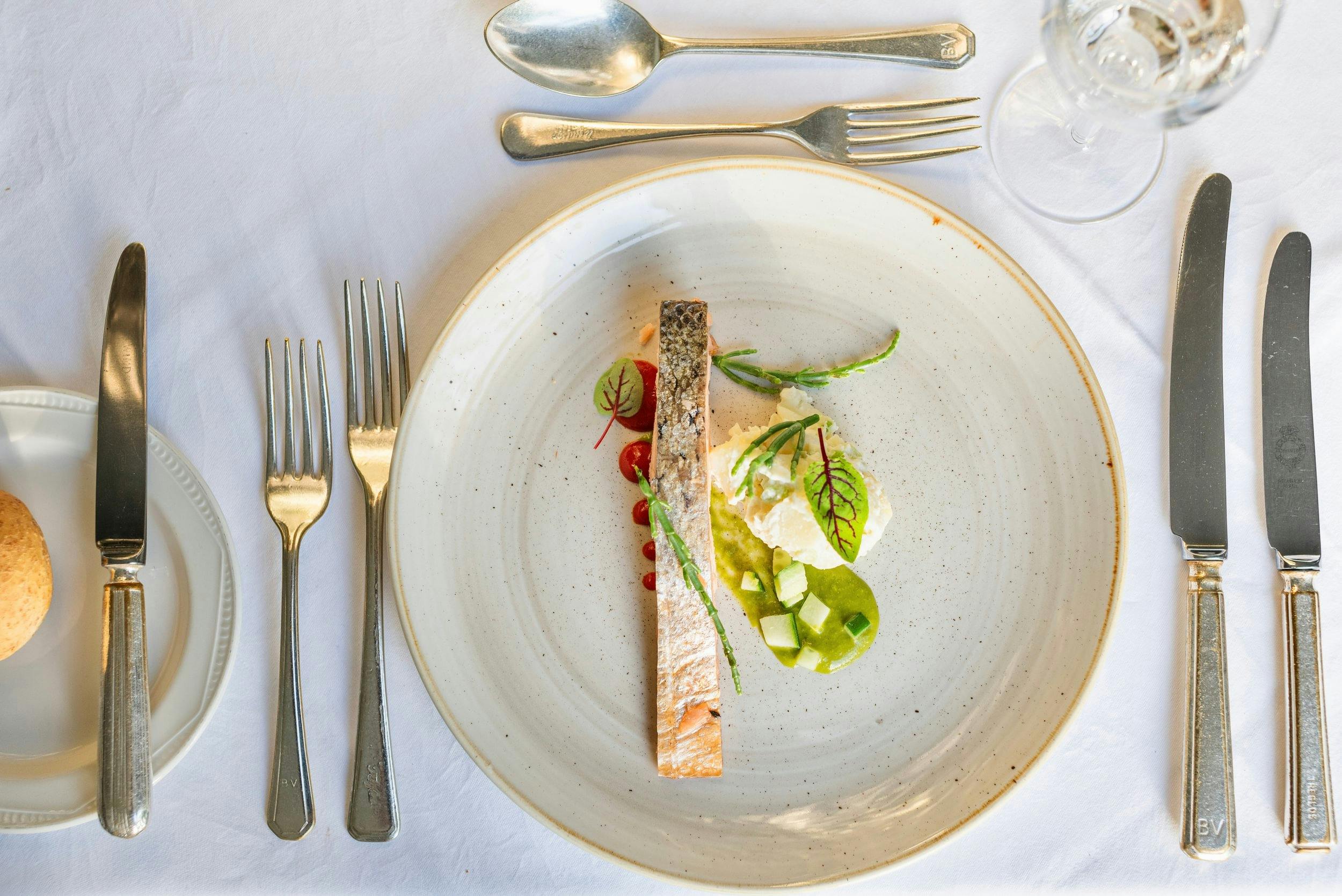 Overhead view of a fine dining place setting with a plated fish dish, silverware, bread plate, and wine glass on a white tablecloth.